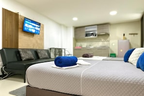 Quarto de hotel moderno com cama, sofá, televisão fixada na parede e área de cozinha compacta.