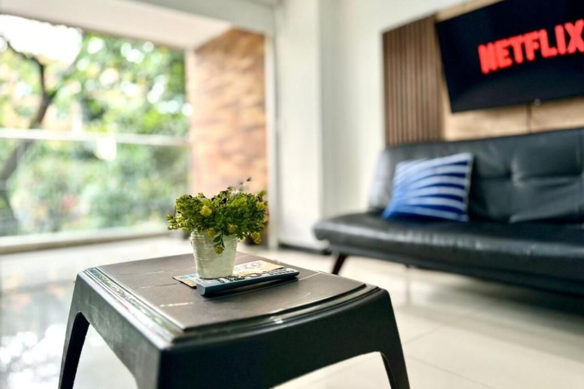 Sala de estar moderna com um sofá preto, uma televisão exibindo 'NETFLIX' e uma mesa pequena com uma planta e controle remoto.