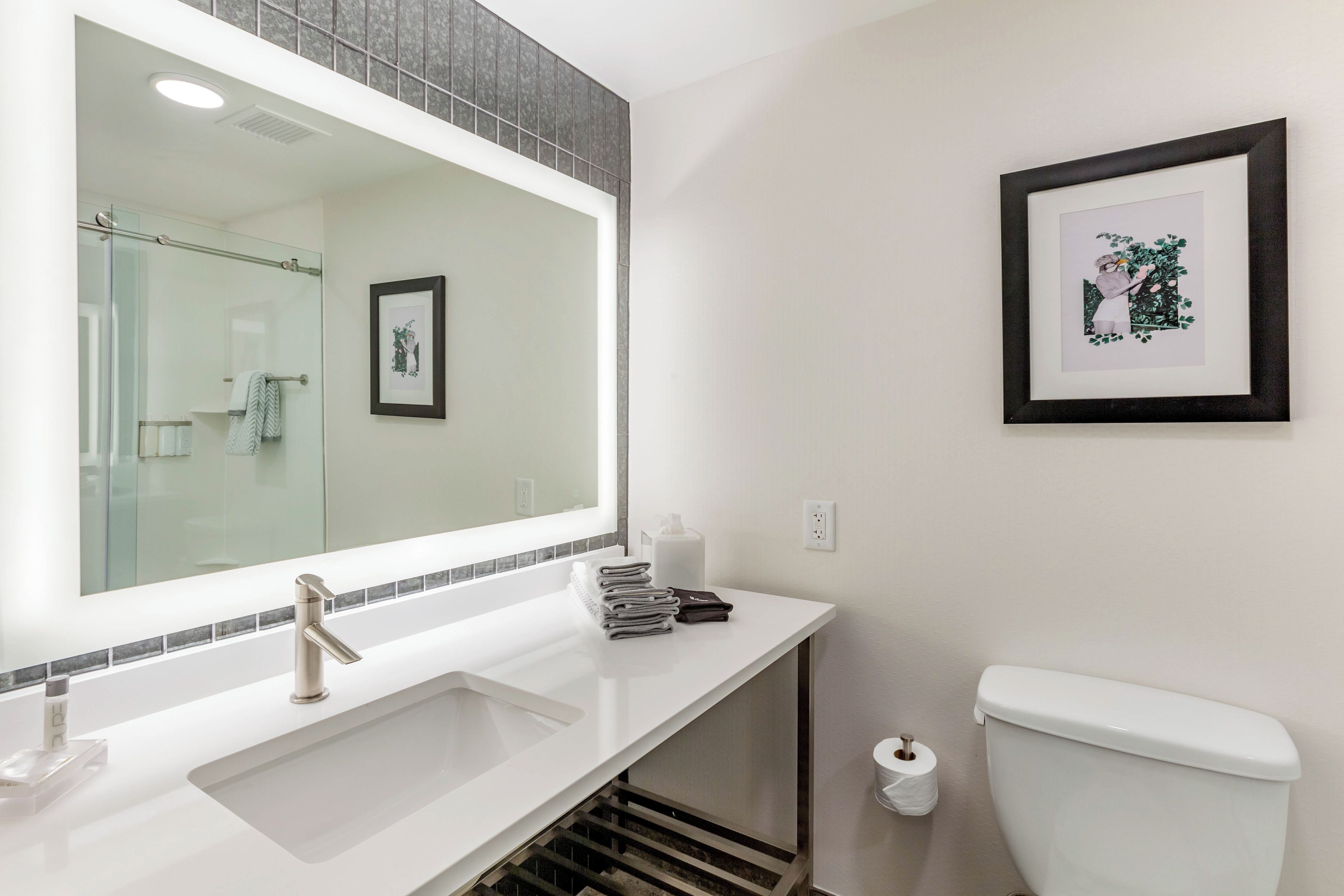 A modern bathroom with a large mirror, a sink, a toilet, and a framed picture on the wall.