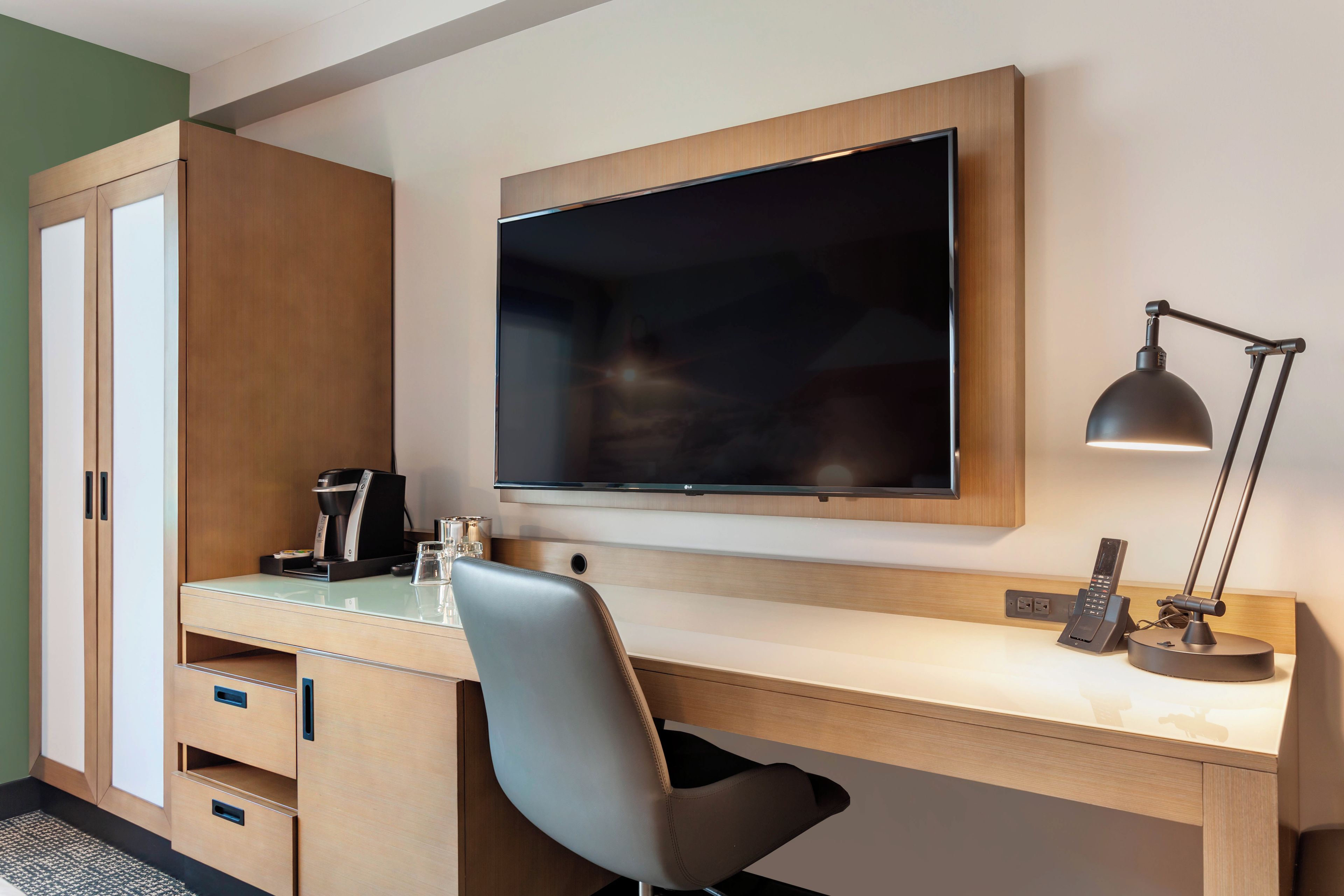 A hotel room with a wooden desk, a flat-screen TV, a desk lamp, a coffee maker, and a chair.