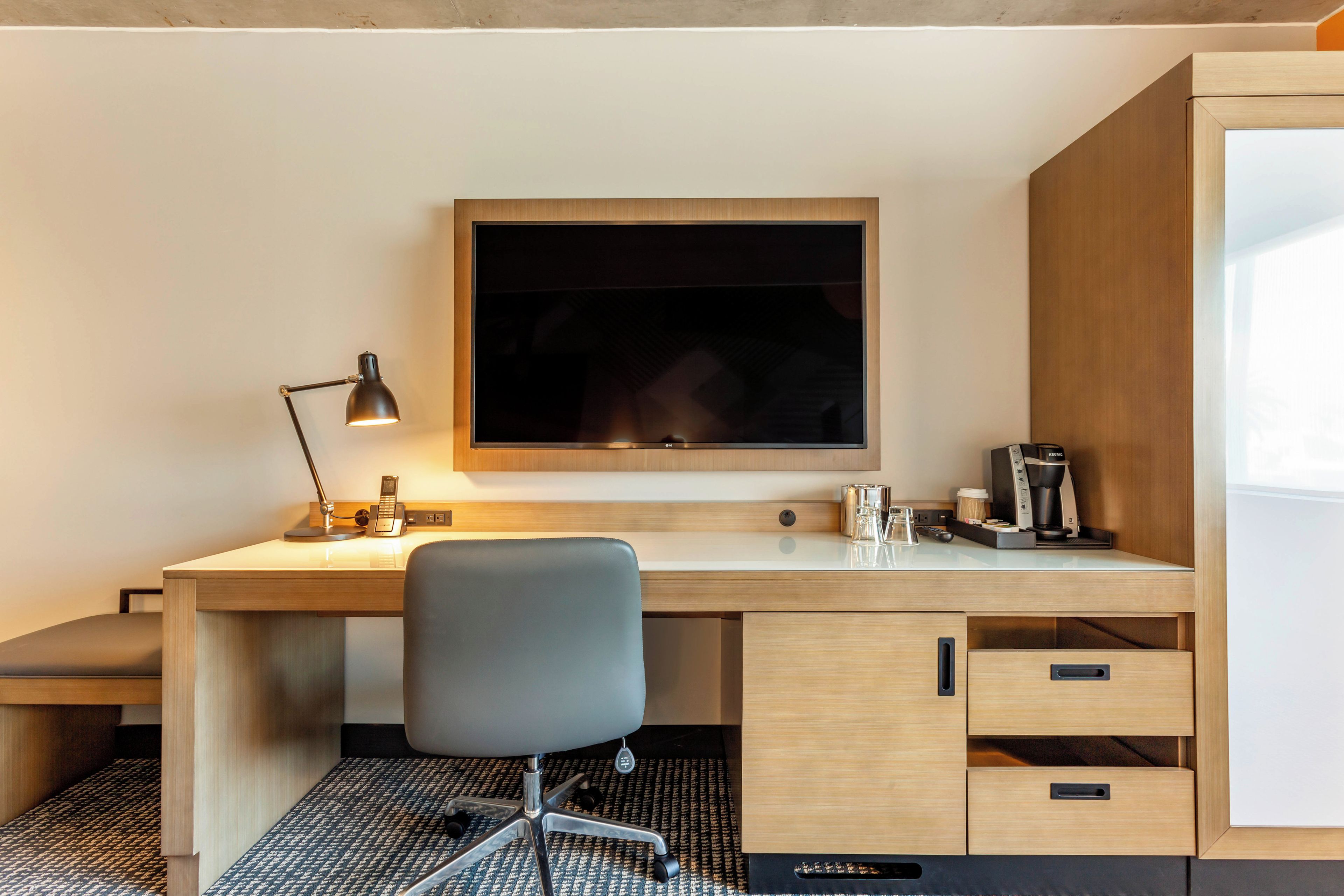 Habitación de hotel con un escritorio de madera, una silla moderna, un televisor de pantalla plana, una cafetera y un teléfono.