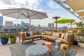 A rooftop terrace with a sectional sofa, two side tables, a patterned ottoman, and umbrellas providing shade.