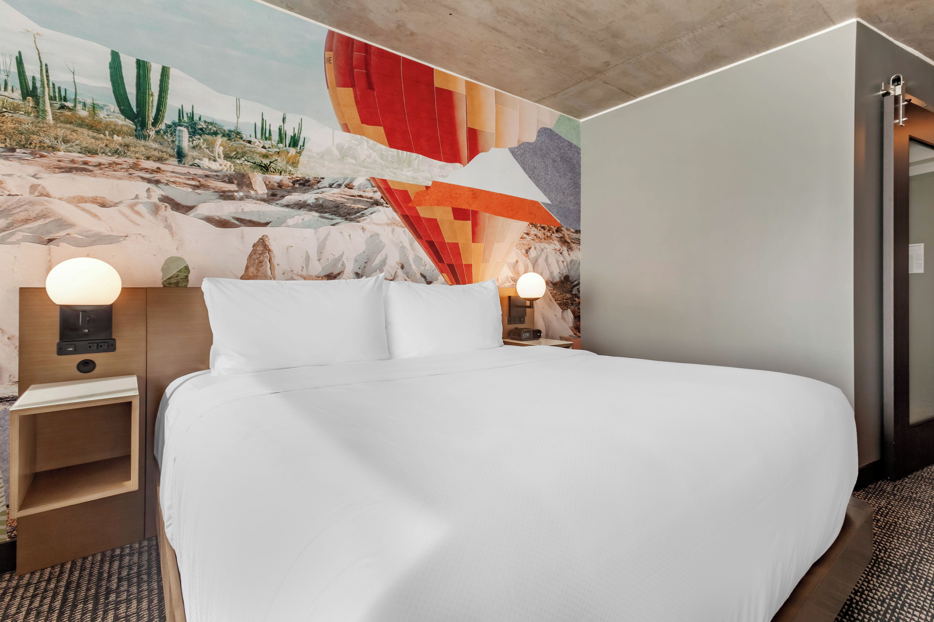 Quarto de hotel com uma cama grande, duas mesas de cabeceira com abajures, um mural na parede e uma porta à direita.