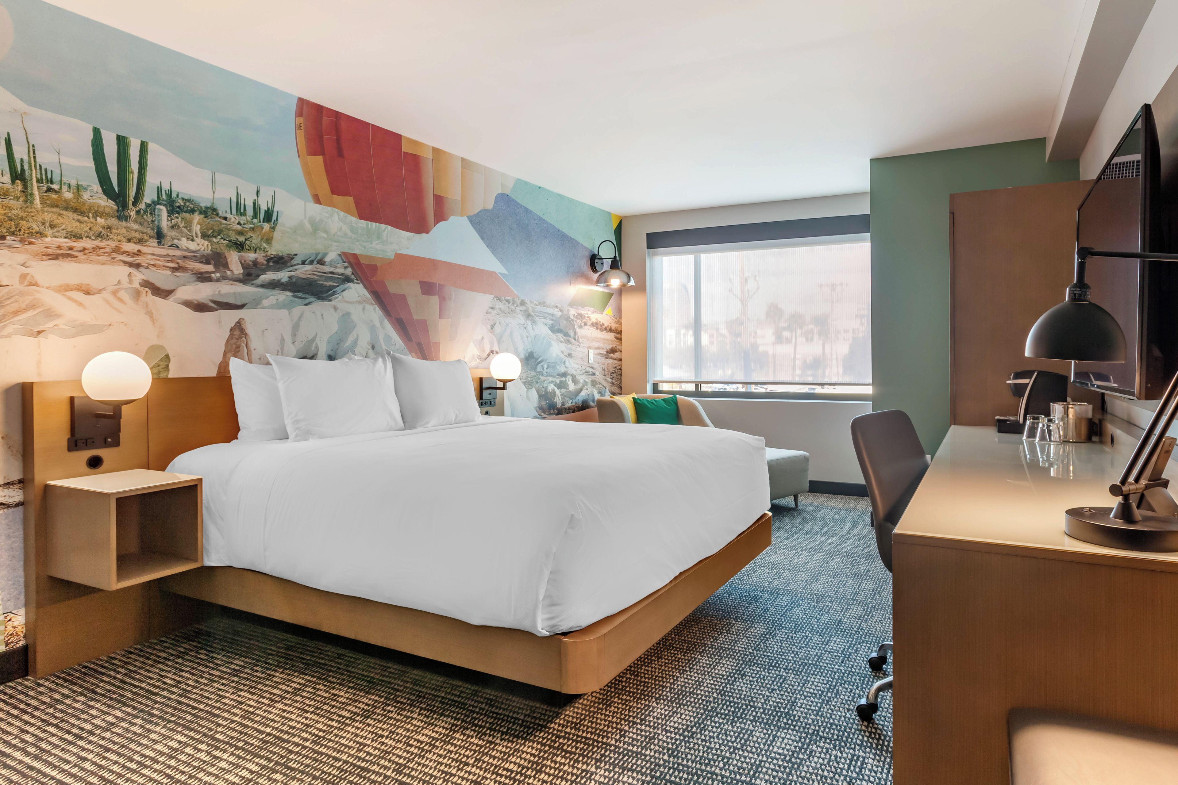 Una habitación de hotel con una cama grande, un escritorio, una silla, una lámpara de escritorio y una pared con un mural abstracto lleno de color.
