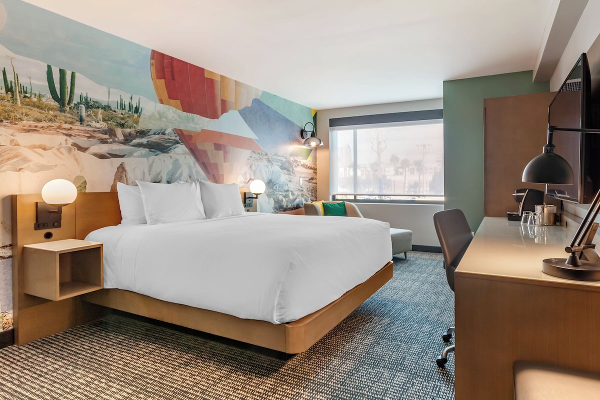 Una habitación de hotel con una cama grande, un escritorio, una silla, una lámpara de escritorio y una pared con un mural abstracto lleno de color.