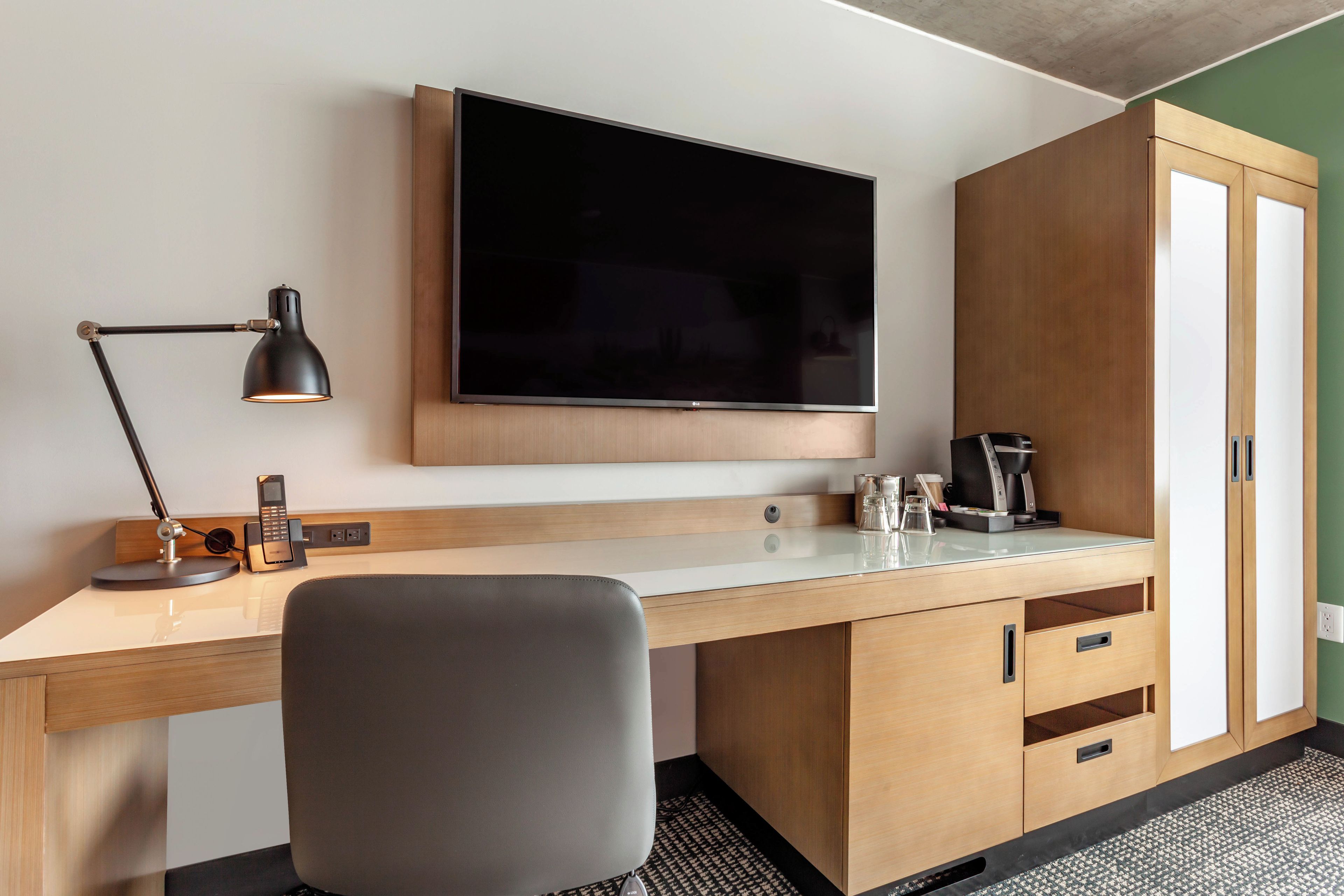 Habitación de hotel con televisor de pantalla plana sobre un escritorio de madera, lámpara de escritorio, una silla, cafetera y minibar.