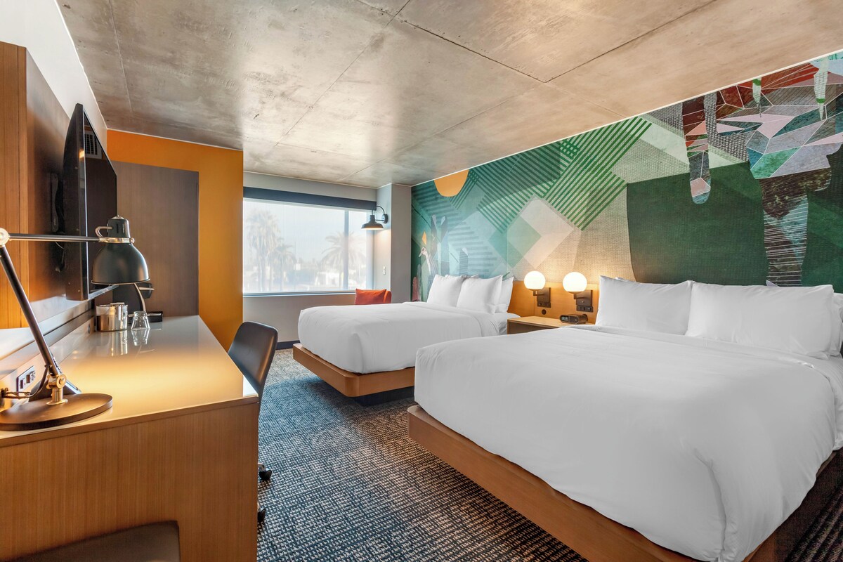 Habitación de hotel con dos camas, un escritorio con lámpara, un ventanal grande y un mural en la pared.
