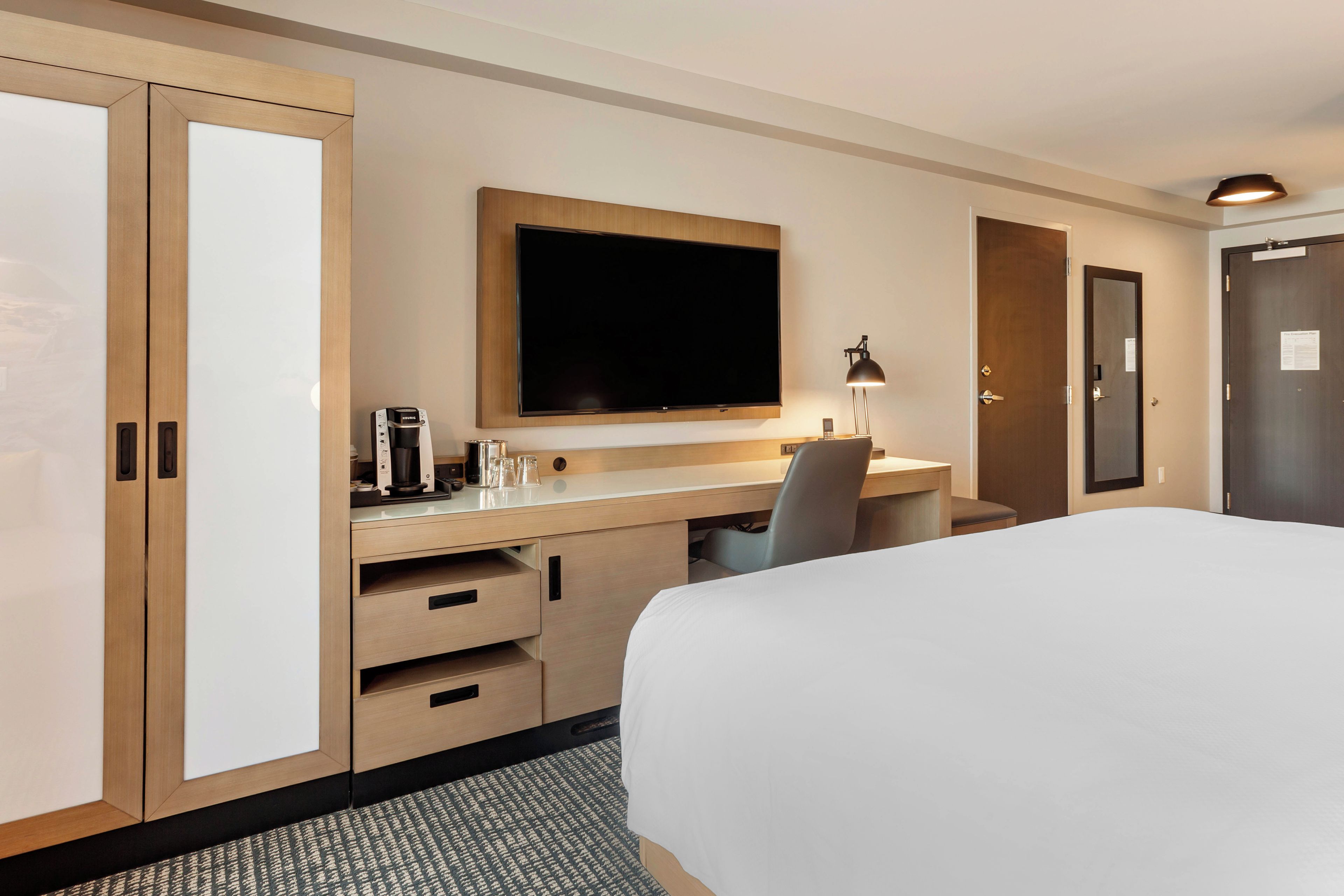 Una habitación de hotel con cama, escritorio, televisor de pantalla plana, cafetera y lámpara.
