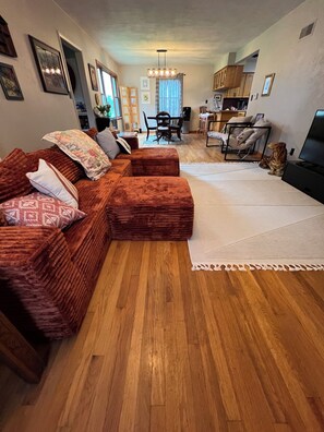 A living room with a sectional sofa, a dining table with chairs, a kitchen area, and a television.