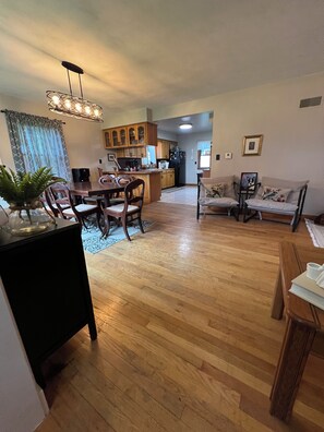 A spacious living area with a wooden dining table, chairs, a sofa, and a kitchen in the background.