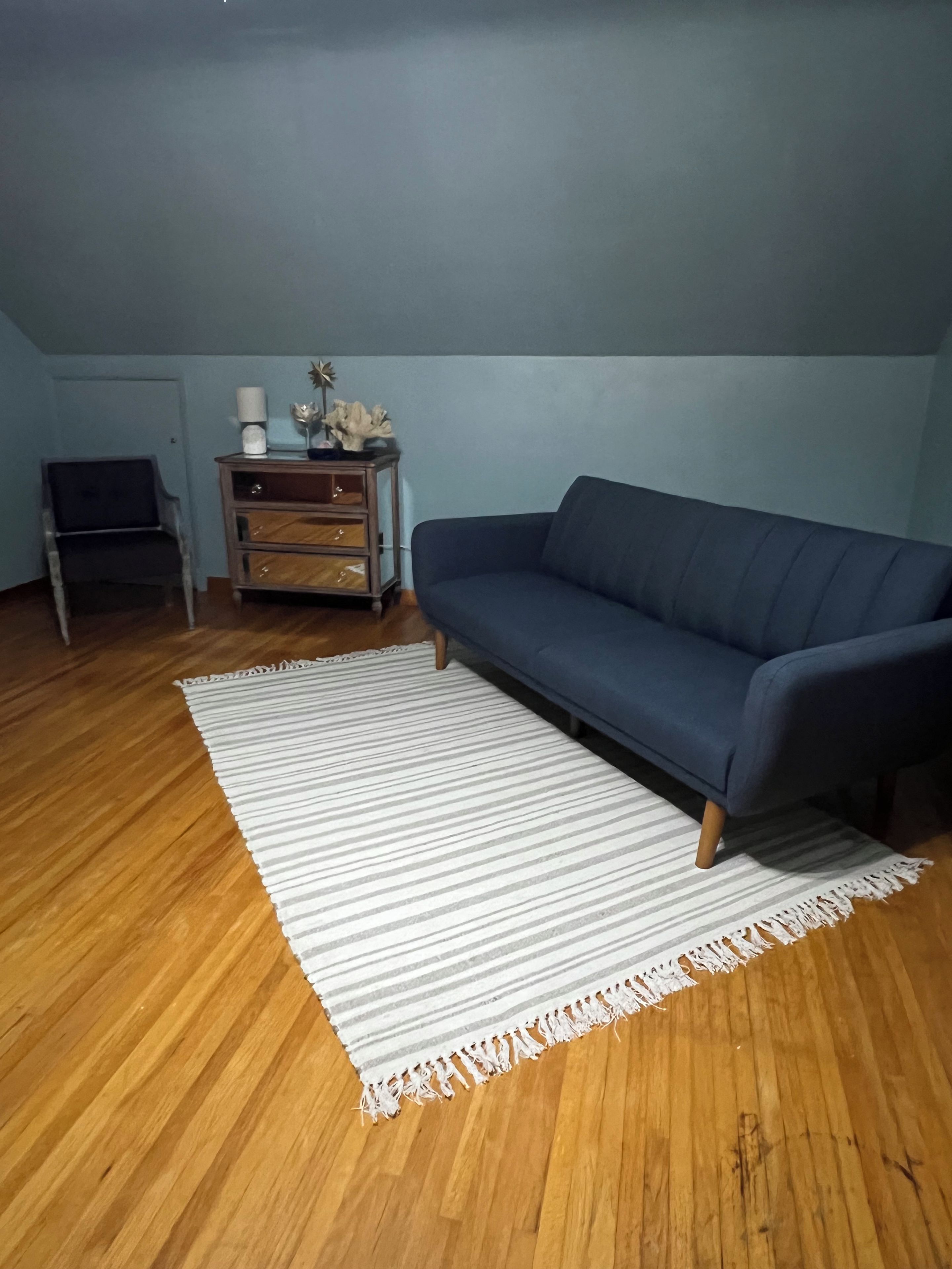 Una habitación con un sofá azul oscuro, piso de madera, alfombra a rayas, un mueble de madera pequeño con objetos decorativos y una silla.
