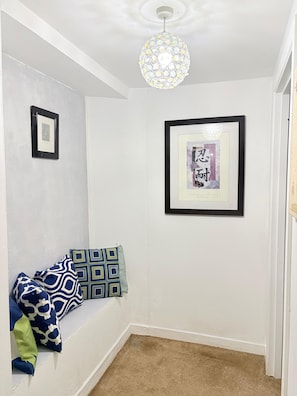 Ein Zimmer mit weißer Wand, einem gerahmten Bild, einem gemusterten Kissen und einer Pendelleuchte.