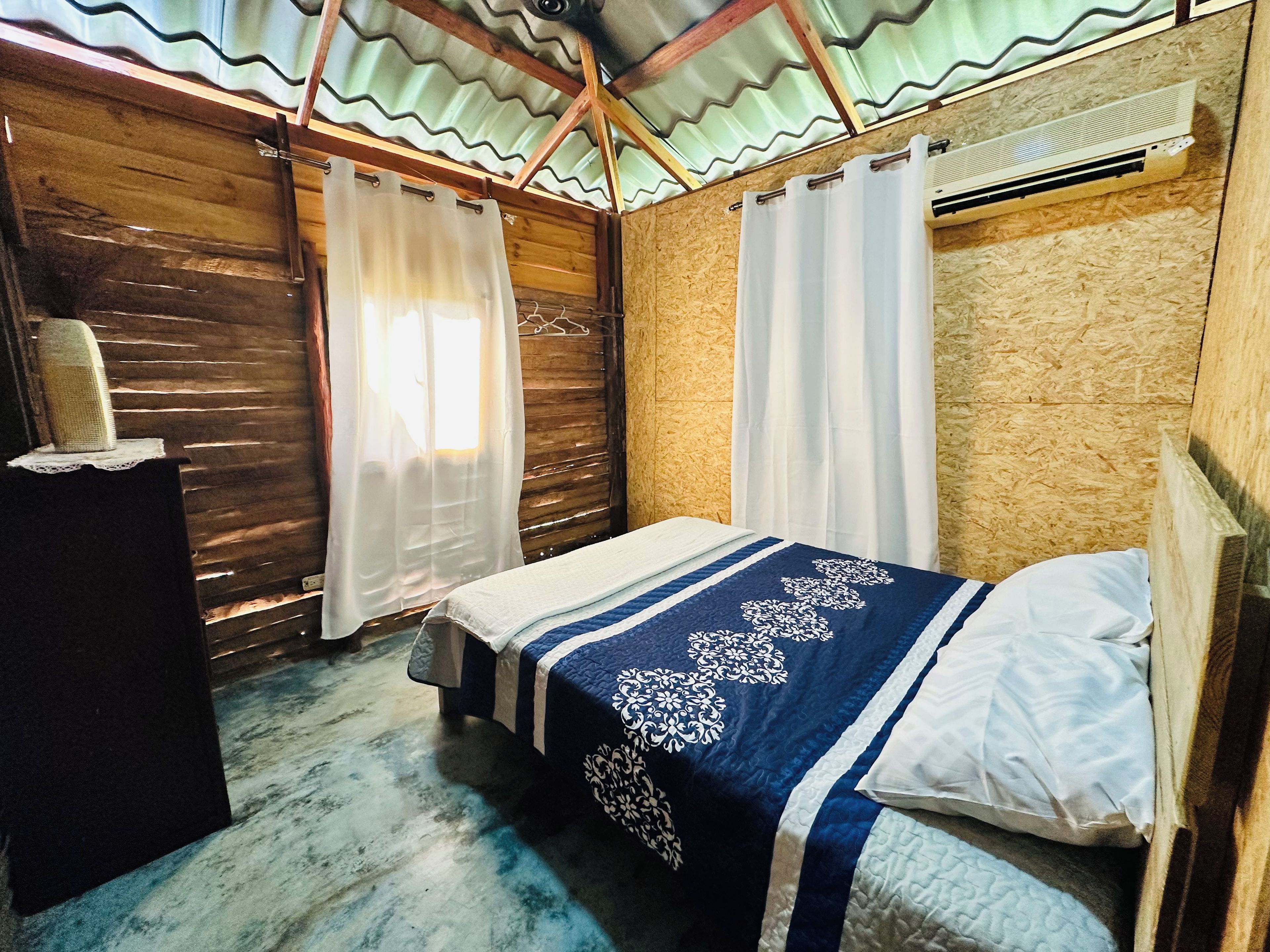 A single bed with a blue and white bedspread, wooden walls, and a corrugated metal roof.