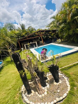 Ein Swimmingpool, umgeben von einem Garten mit einer Pergola, Palmen und einer Rasenfläche.