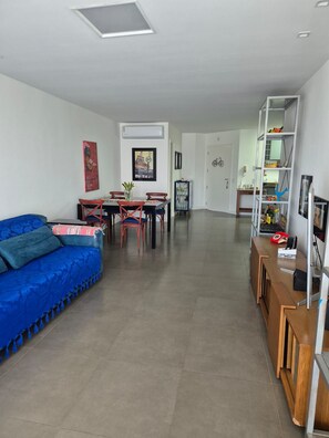 A modern living room with a blue sofa, dining table, and chairs. There is a kitchen area with a white cabinet and a bicycle. - Apto 2 quartos em resort a passos da praia – Cachoeira do Bom Jesus (Florianópolis)