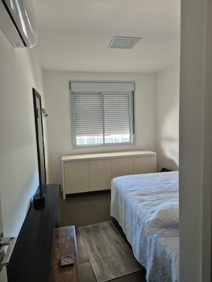 Ein Schlafzimmer mit einem Bett, einem Fenster mit Jalousien, einem Heizkörper und einem Schreibtisch mit Lampe.