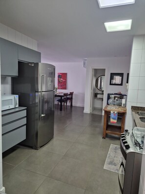 A kitchen with a refrigerator, microwave, and a dining area with chairs and a table. - Apto 2 quartos em resort a passos da praia – Cachoeira do Bom Jesus (Florianópolis)