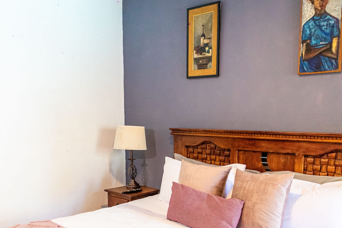 Quarto de hotel com uma cama, uma mesa de cabeceira, um abajur e duas pinturas na parede.
