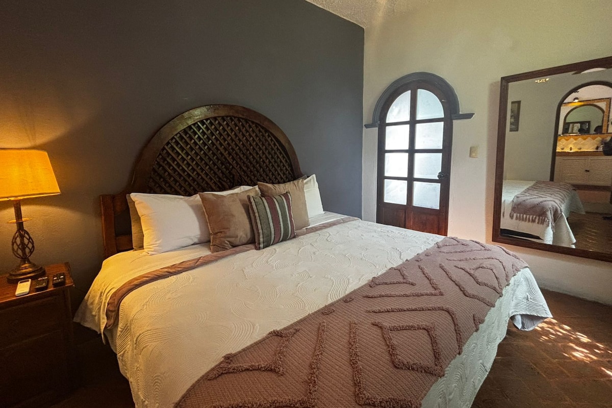 Uma cama bem arrumada com cabeceira de madeira, luminária de cabeceira e um espelho grande na parede.
