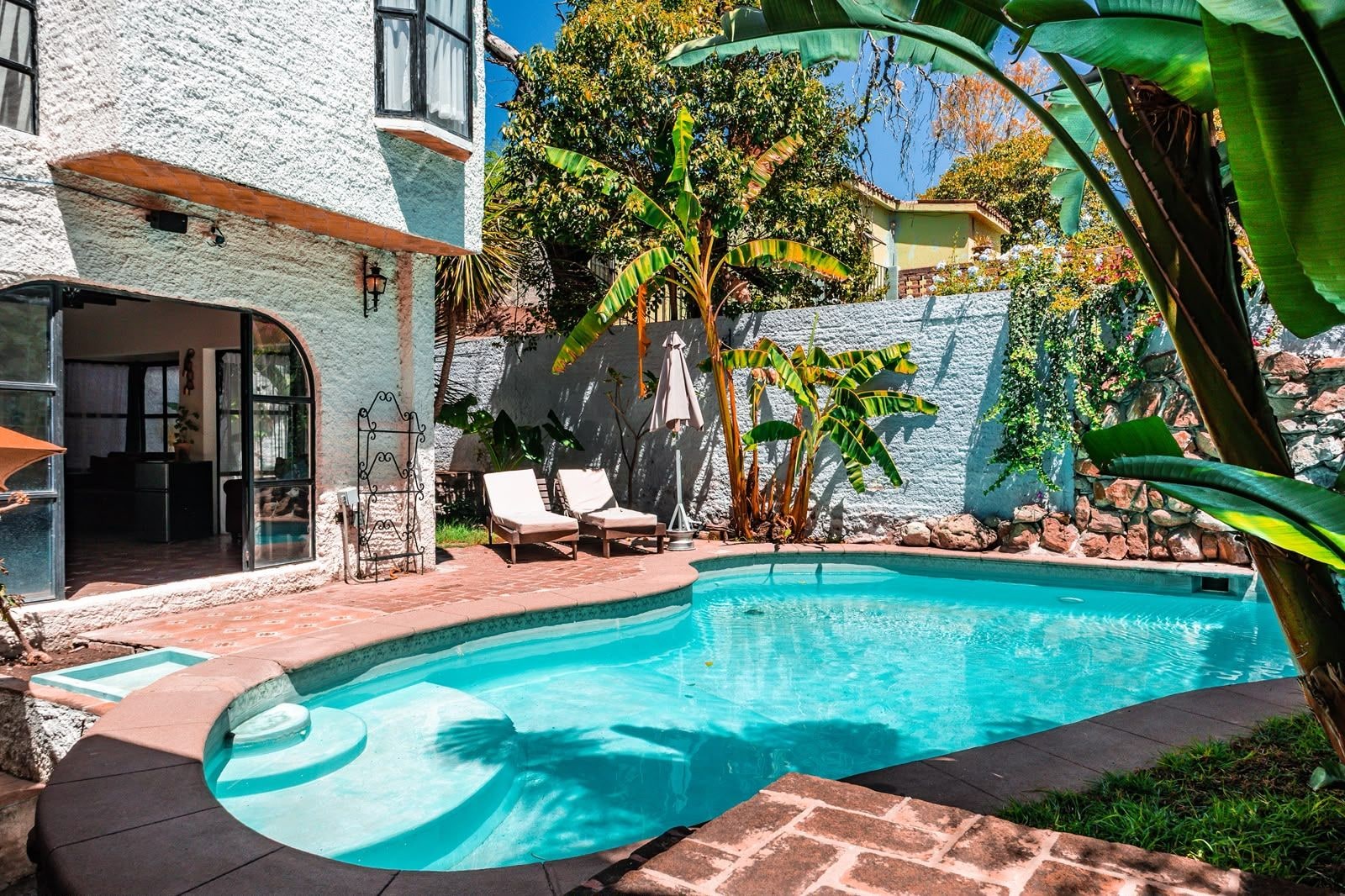 A swimming pool with lounge chairs, a stone wall, and tropical plants.