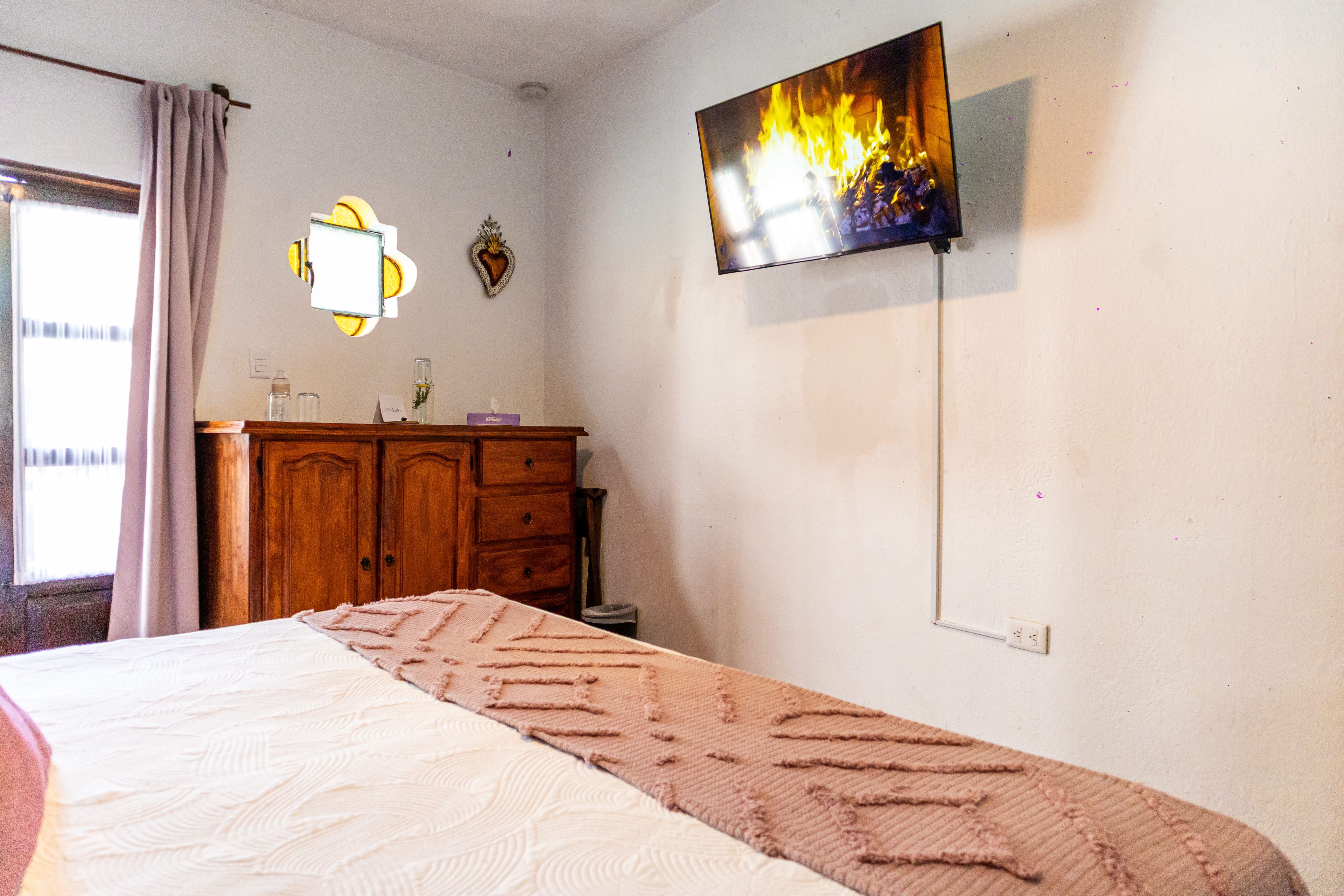 Une chambre à coucher avec un lit, un téléviseur fixé au mur, une commode et une fenêtre avec des rideaux.