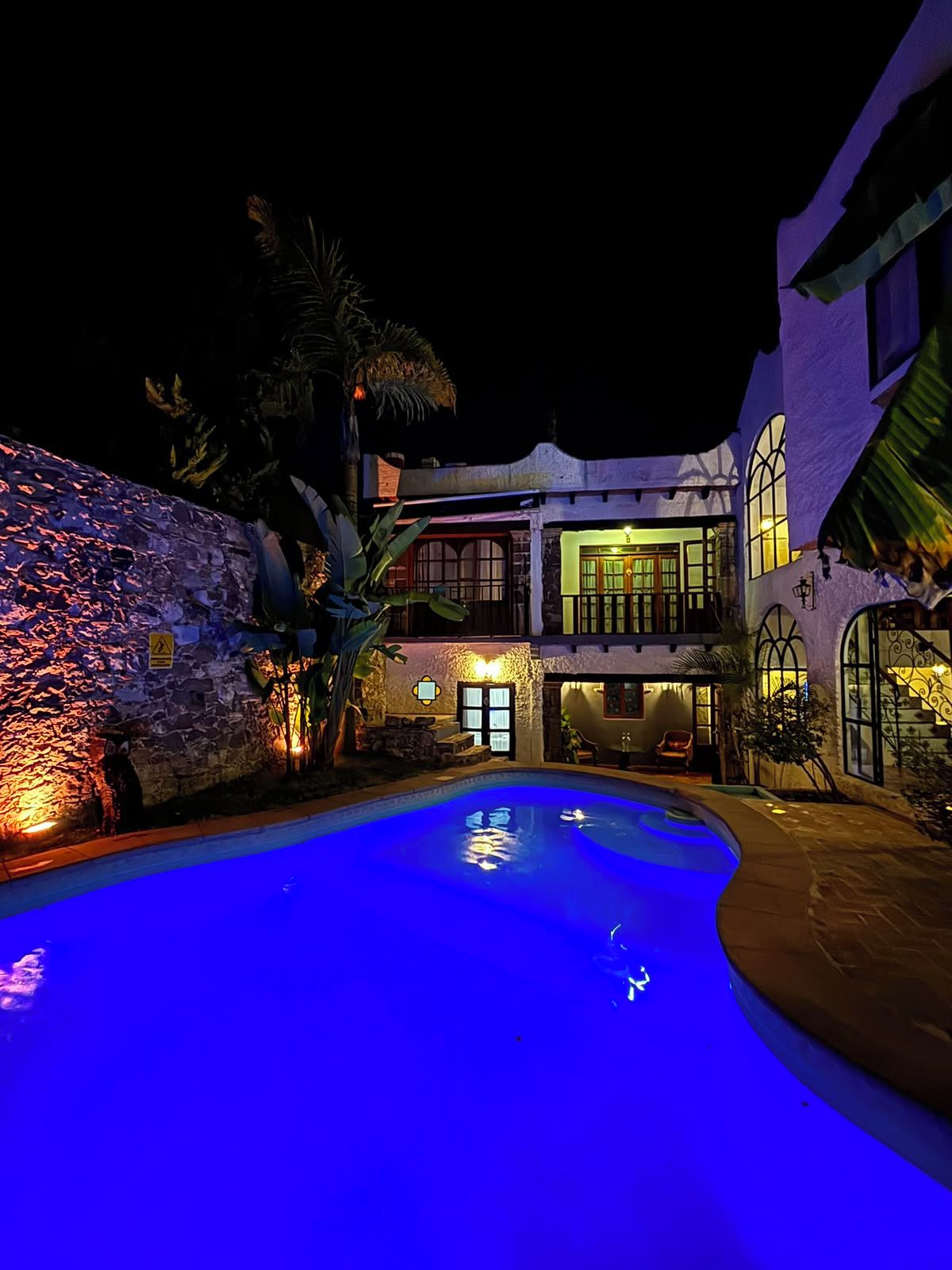 Vista nocturna de un resort con piscina, pared de piedra y un edificio de dos plantas con ventanas iluminadas.