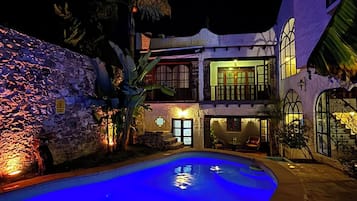Vista nocturna de un resort con piscina, pared de piedra y un edificio de dos plantas con ventanas iluminadas.