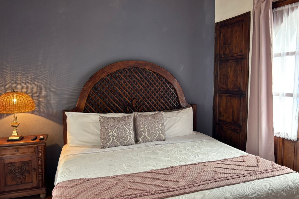 Quarto com uma cama grande, cabeceira de madeira, mesa de cabeceira com abajur e janela com cortinas.