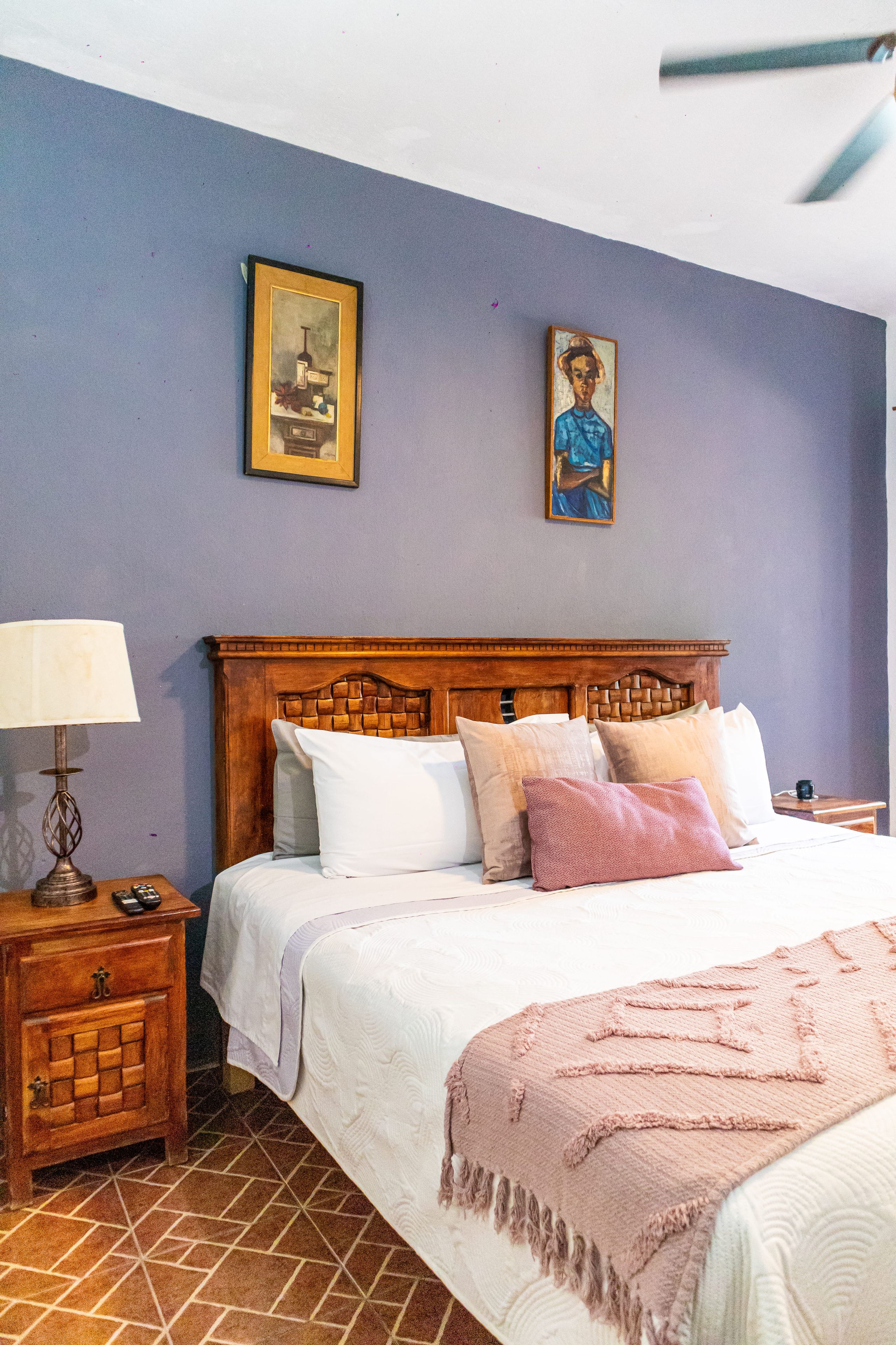 A bedroom with a wooden headboard, a bed with multiple pillows, a nightstand with a lamp, and two framed pictures on the wall.