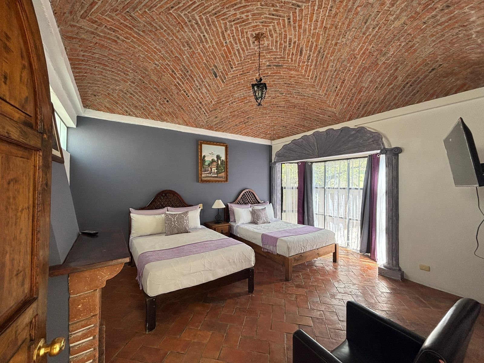 A room with two beds, a wooden door, a brick ceiling, a painting, and a window with curtains.