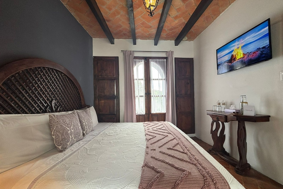 Quarto com cama, cabeceira de madeira, mesa de cabeceira com luminária, pintura na parede e janela com cortinas.