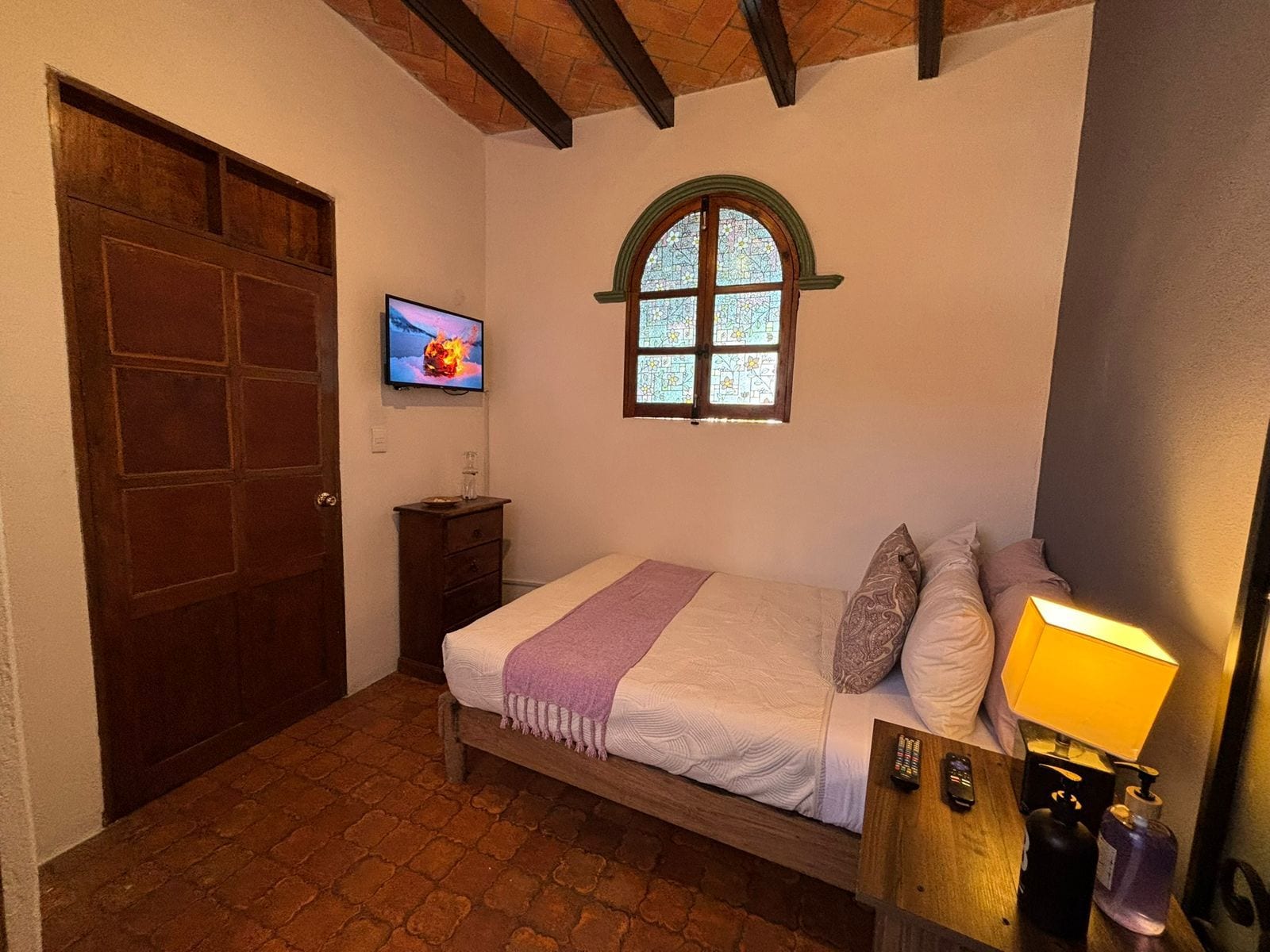 Un dormitorio con una cama, un televisor montado en la pared, un ventanal con marco verde y una puerta de madera.