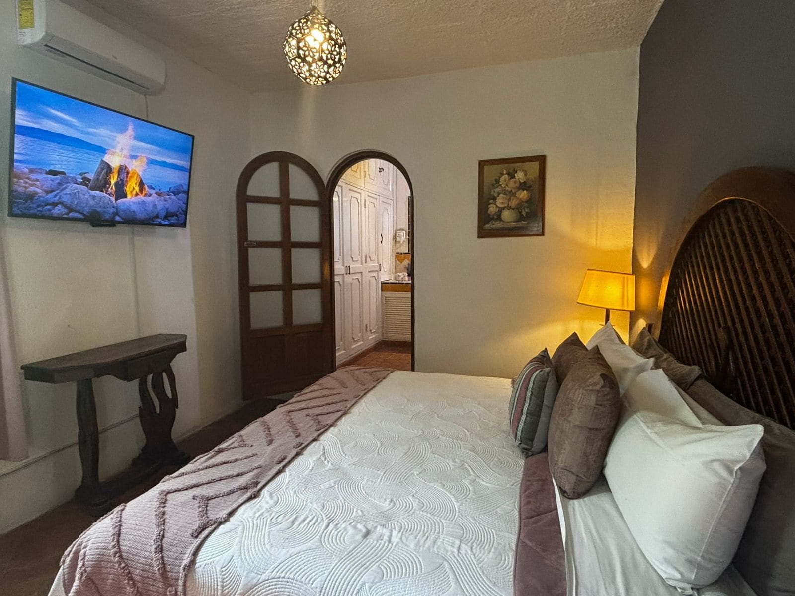 A bedroom with a bed, a TV, a lamp, and a door leading to a kitchen.