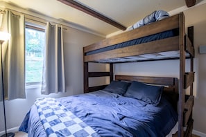 Een stapelbed met een geruit sprei, een raam met gordijnen en een plafond van houten balken.