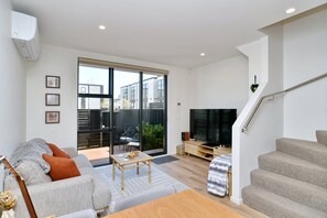 A modern living room with a sofa, coffee table, and TV. There is a staircase and a sliding glass door leading to an outdoor area. - City-side Riccarton, Walk to Mall - Matai St West (Riccarton)