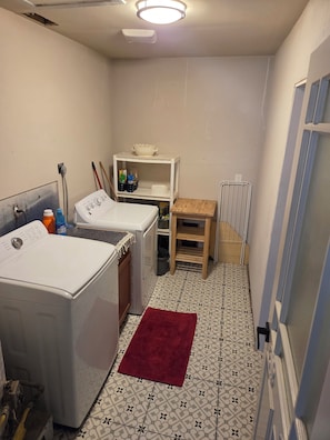 A laundry room with two washing machines, a shelf with cleaning supplies, a small table, and a radiator. - Modern cabin style home in the heart of San Antonio. (San Antonio)