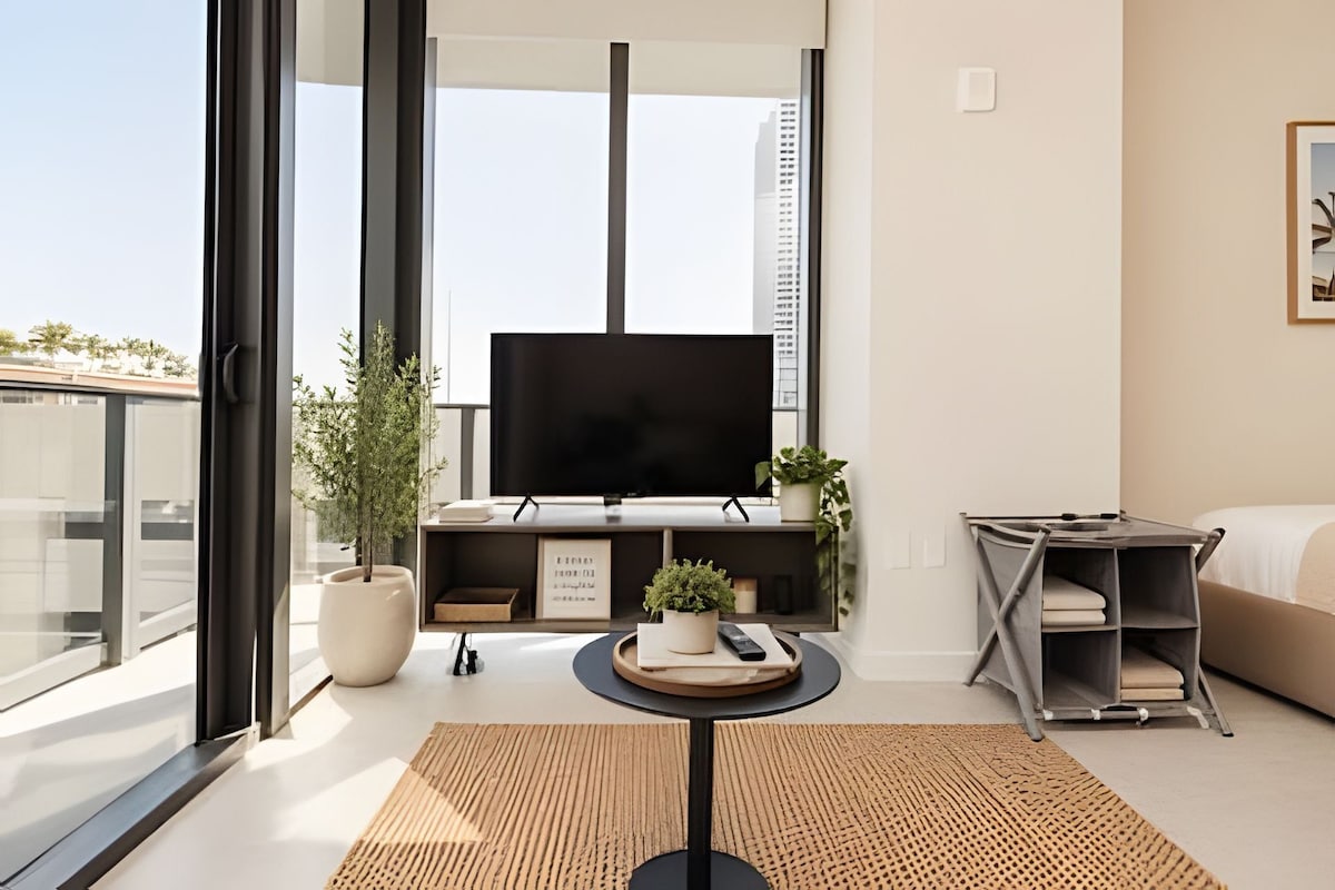 Uma sala de estar moderna com uma televisão grande, um sofá, uma mesa de centro e uma varanda com vista para edifícios.