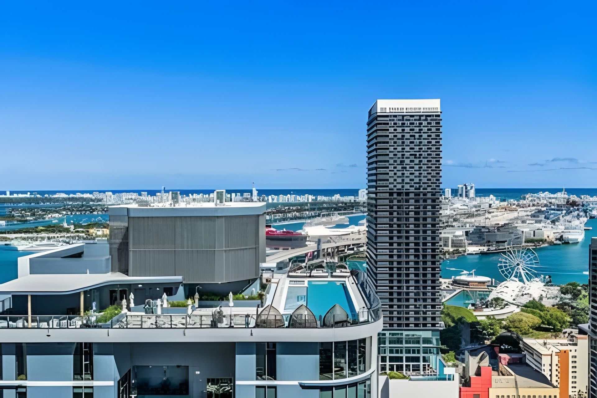 Um edifício moderno e imponente, com design angular característico, localizado em uma paisagem urbana costeira com um porto de iates e uma grande roda-gigante ao fundo.