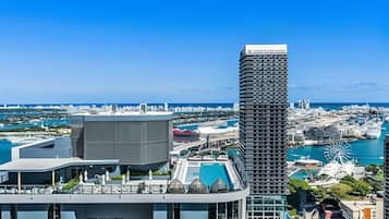Um edifício moderno e imponente, com design angular característico, localizado em uma paisagem urbana costeira com um porto de iates e uma grande roda-gigante ao fundo.