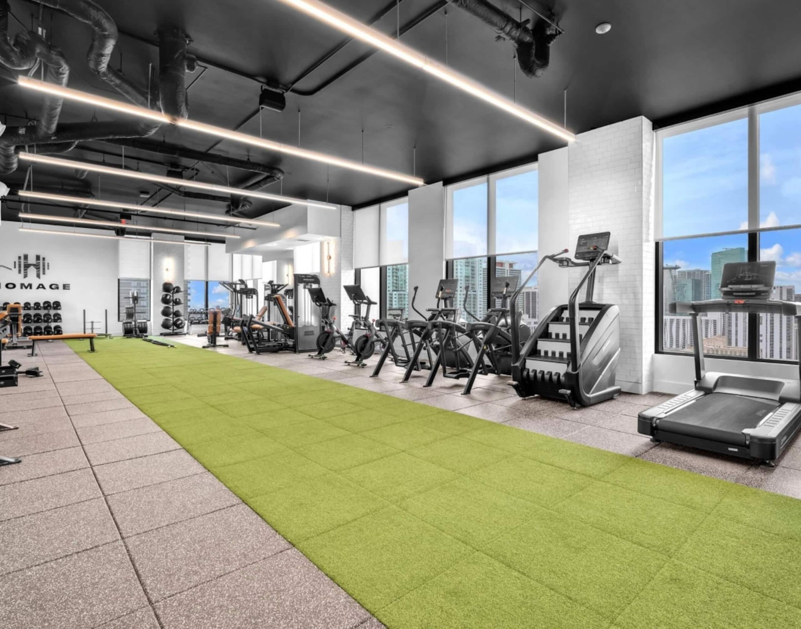 Un gimnasio con equipo de ejercicio, una zona de césped artificial y amplios ventanales que ofrecen vistas a la ciudad.