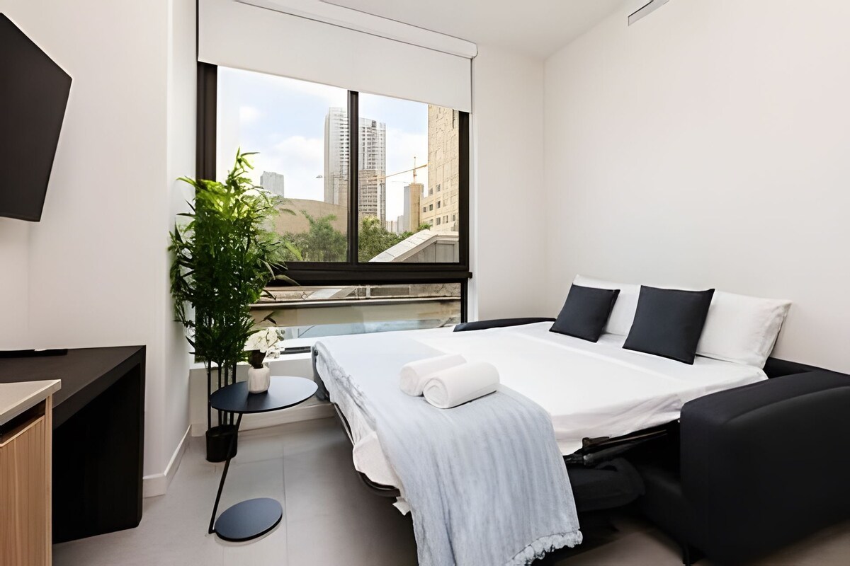Quarto de hotel moderno com cama, escrivaninha, poltrona, planta em vaso e vista para a paisagem urbana.