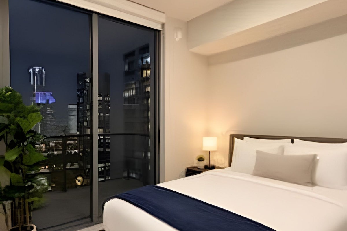 Quarto de hotel com cama grande, mesa de cabeceira, luminária, planta em vaso e vista da cidade à noite.