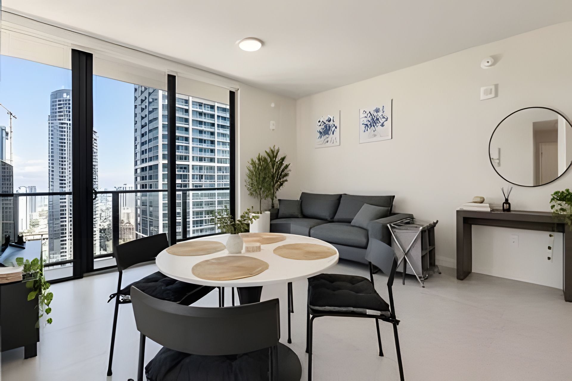 A modern living room with a round table, grey sofa, and large windows offering a city view.