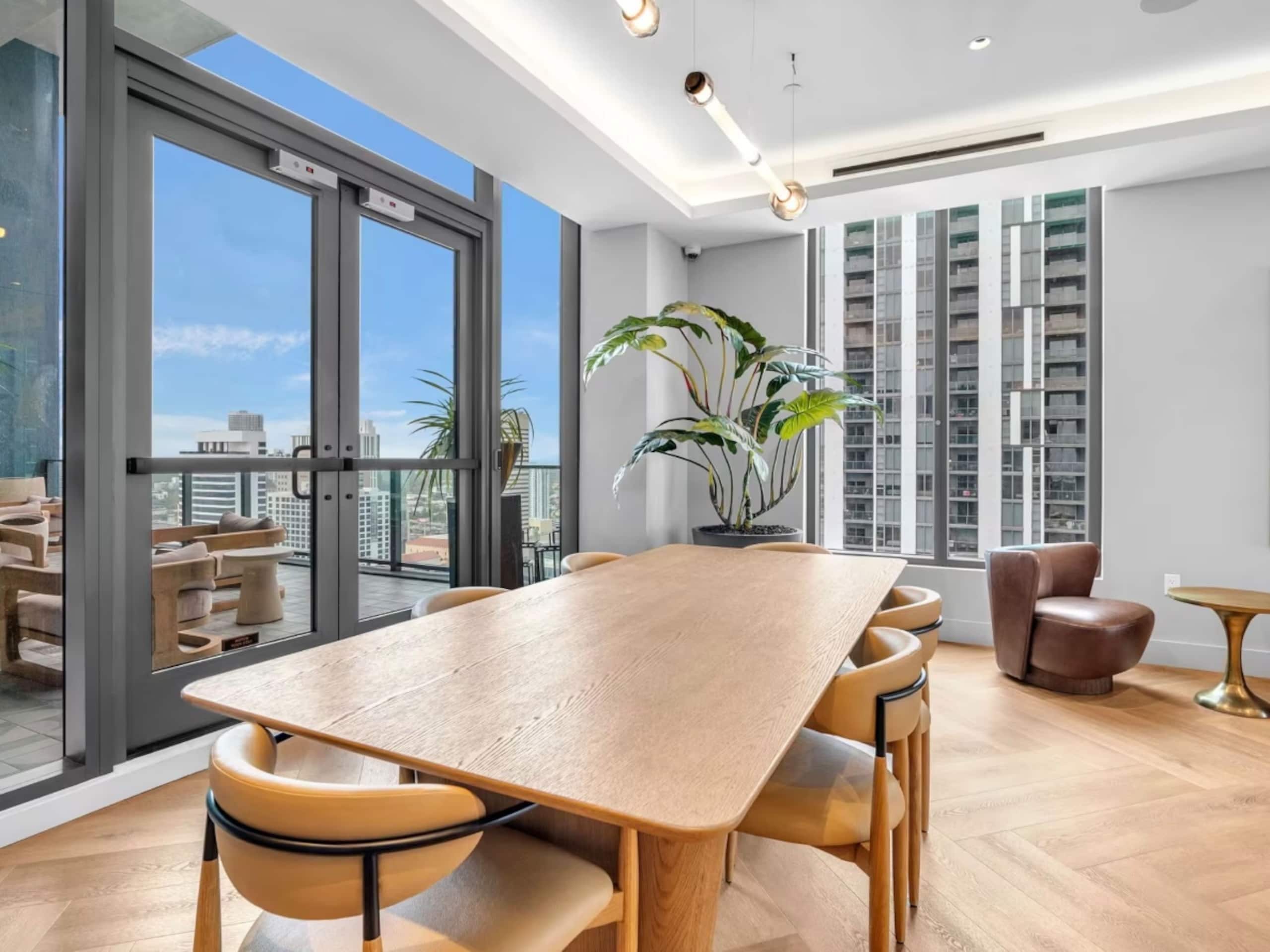 A modern interior with a large wooden table, comfortable seating, a potted plant, and a view of the city through large windows.