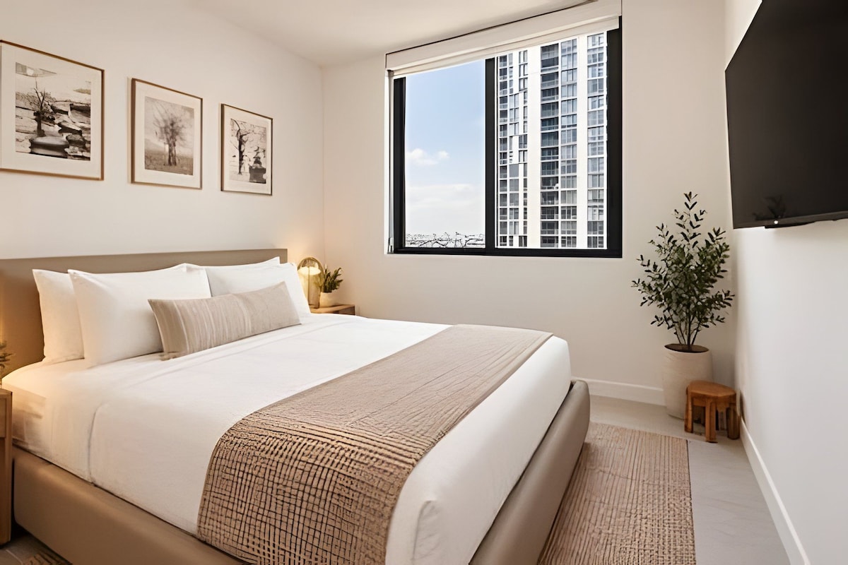 Quarto com uma cama grande, janela com vista para edifícios e três quadros na parede.