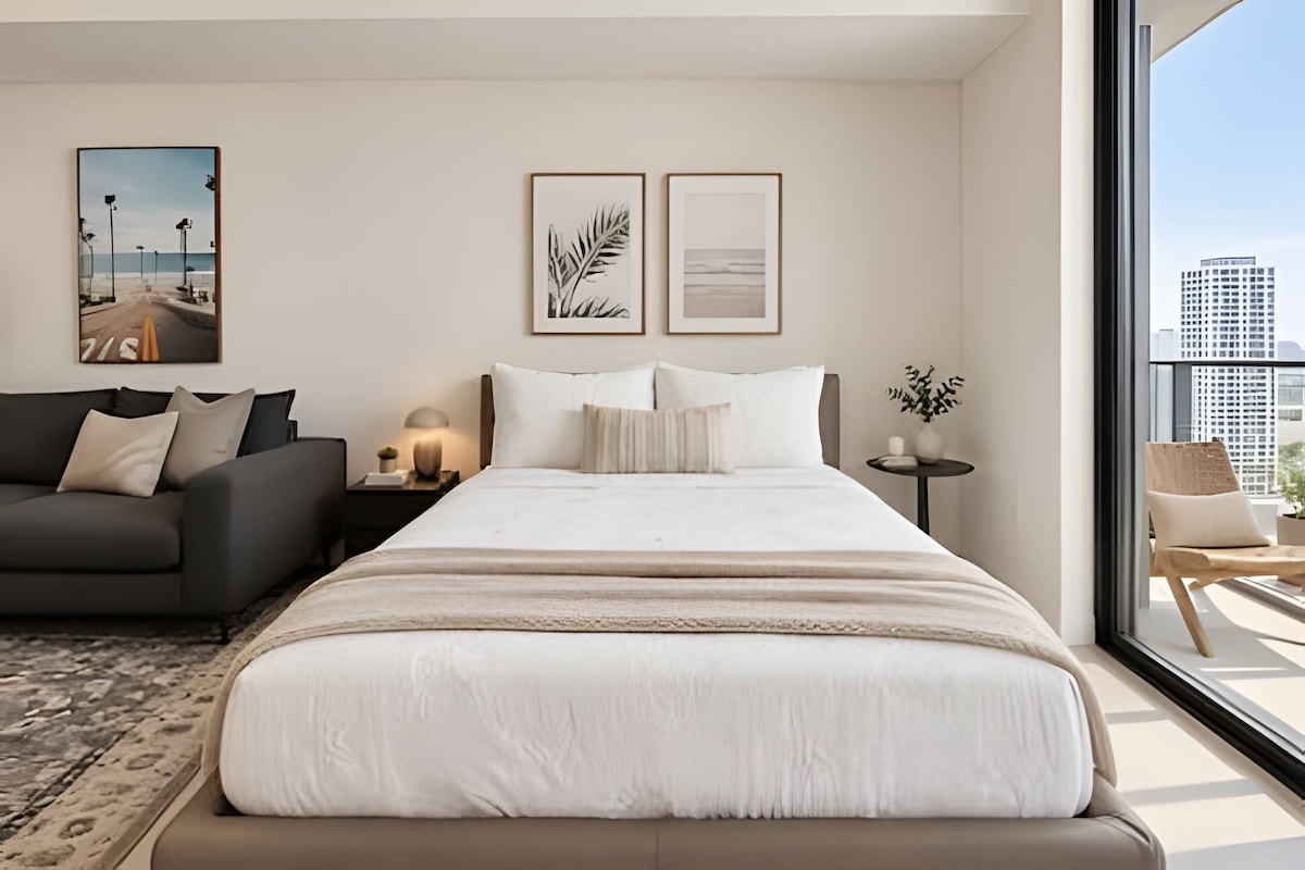 Una habitación de hotel moderna con una cama grande, un sofá, una mesita con lámpara y vistas a la ciudad.