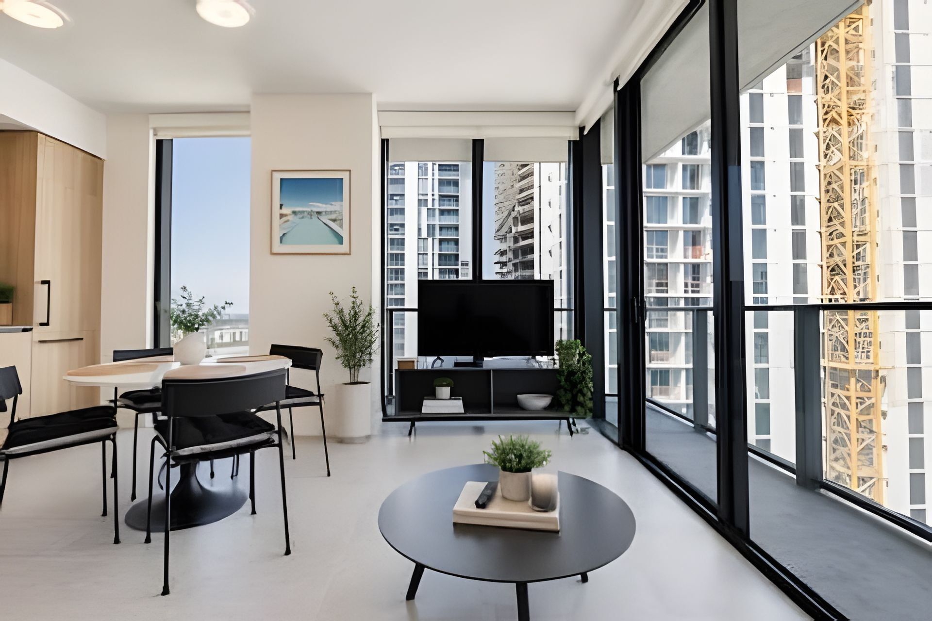 A modern living room with a dining area, a flat-screen TV, and large windows offering a view of high-rise buildings.