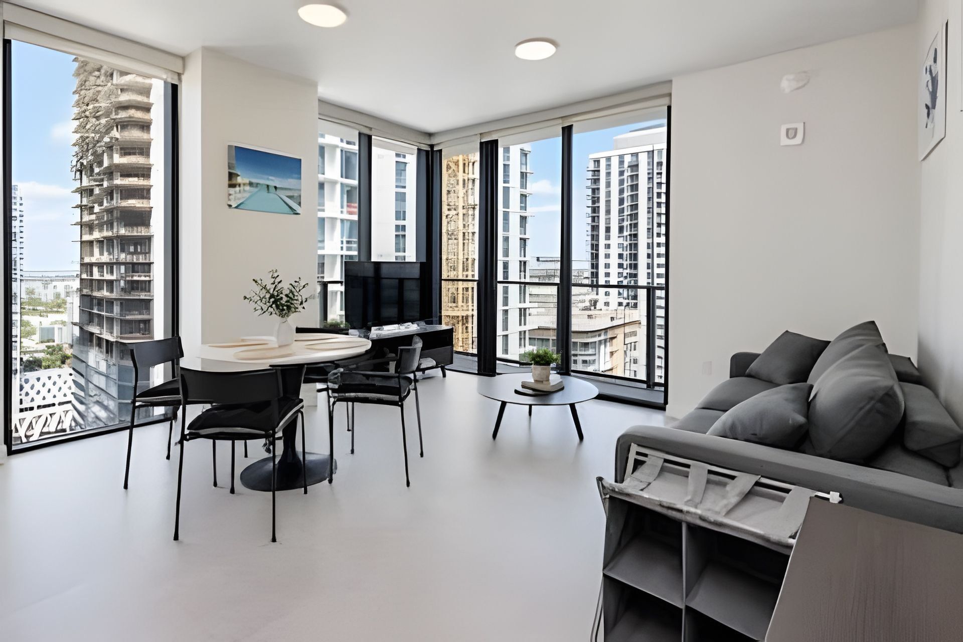 A modern living room with a sofa, a dining table with chairs, a TV, and large windows offering a view of surrounding buildings.
