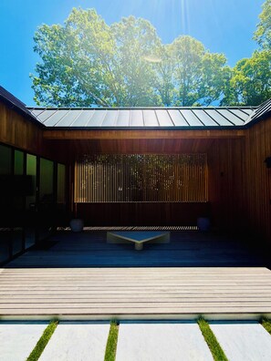 A modern outdoor space with a wooden deck, seating area, and a view of trees. - Brand new 2026 designer construction- Full privacy-Gym-Sauna-Hotel bar- Firepit. (East Hampton)