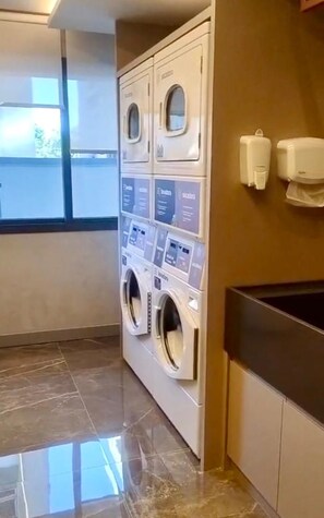 A laundry area with stacked washers and dryers, a tiled floor, and a window providing natural light. - New studio apartment in Cidade Baixa, near Redenção Park and UFRGS (Federal University of Rio Grande do Sul). (Porto Alegre)